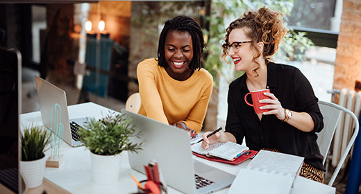 Two women sitting at a table online shopping on a laptop One women is laughing and holding a coffee mug